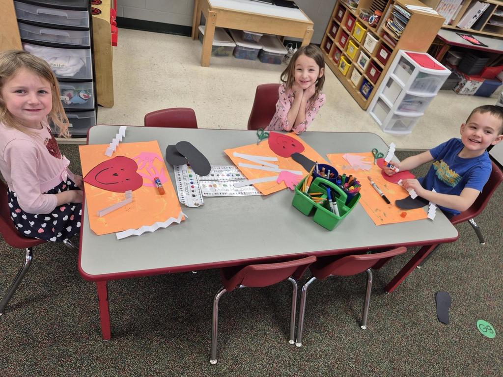Children seated at a table working on heart-themed crafts with paper cutouts, glue sticks, and markers.