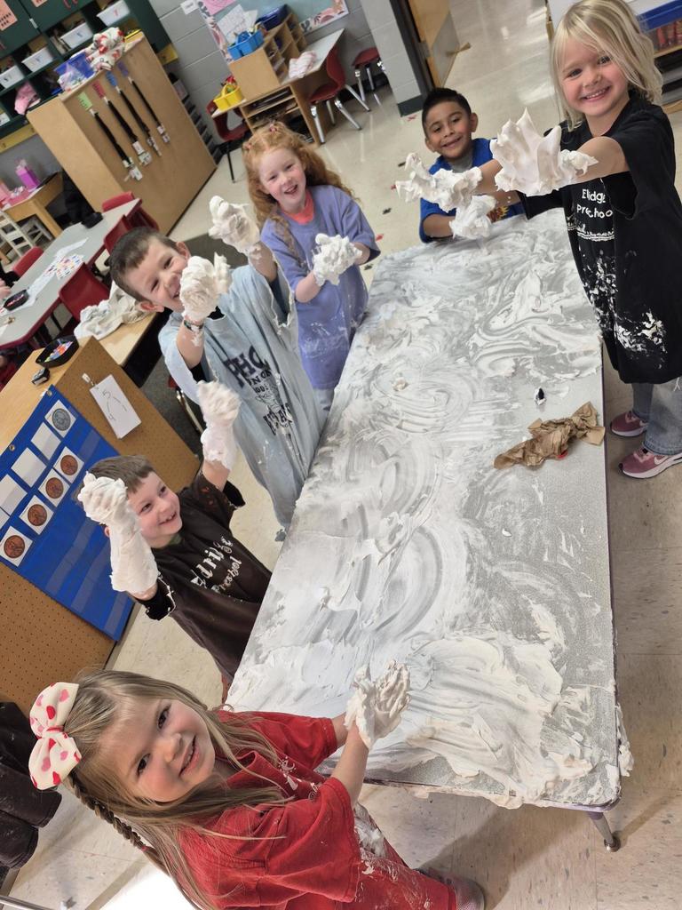 Students gathered around a table covered in shaving cream, grinning and showing their hands during a hands-on classroom activity.