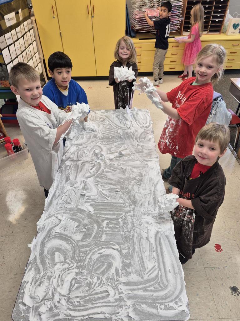 Young students standing around a table covered in shaving cream, smiling and holding up their messy hands during a sensory activity.