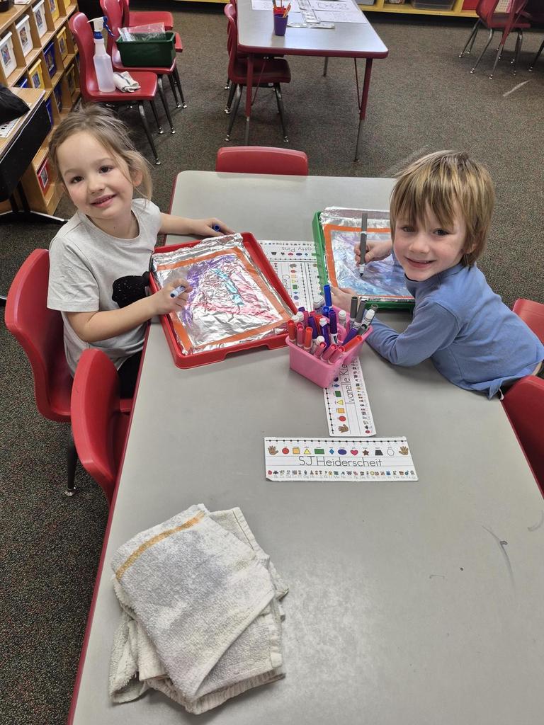 Two young students drawing with markers on foil-covered trays during a creative classroom activity.