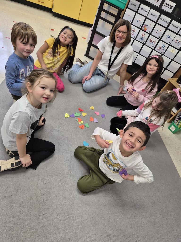 Teacher sitting on the classroom carpet with young students holding colorful paper hearts during a small group activity.