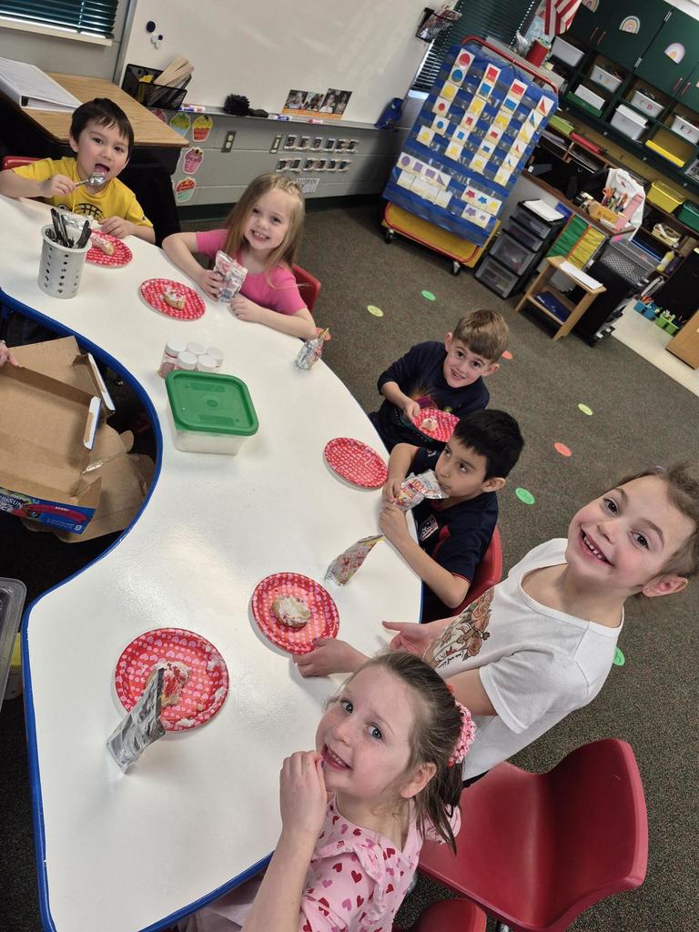 Kindergarten students sitting at a kidney-shaped table enjoying frosted cookies and juice boxes during a classroom celebration.