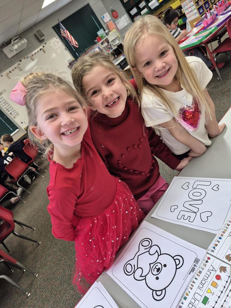 Three young girls smiling at a classroom table with Valentine-themed coloring pages featuring hearts and a teddy bear.