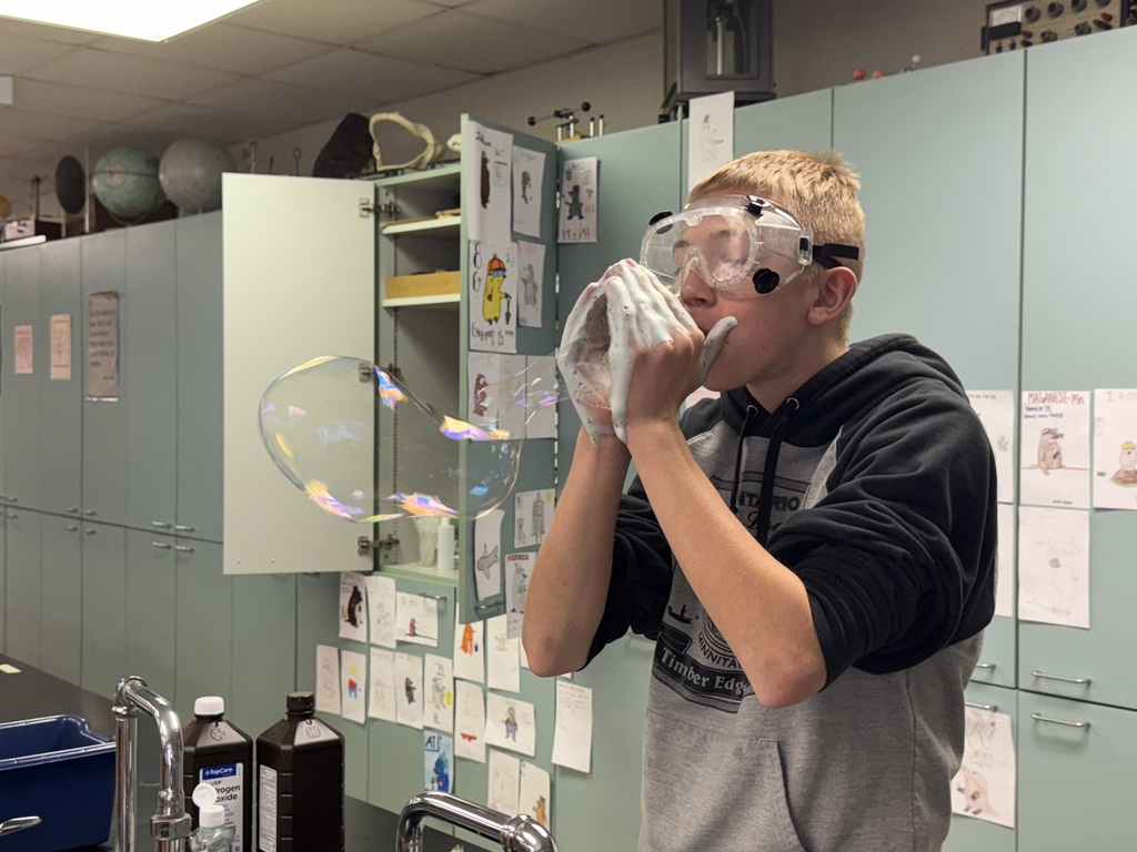 Student in safety goggles blows a large iridescent bubble formed from a foamy chemistry experiment.