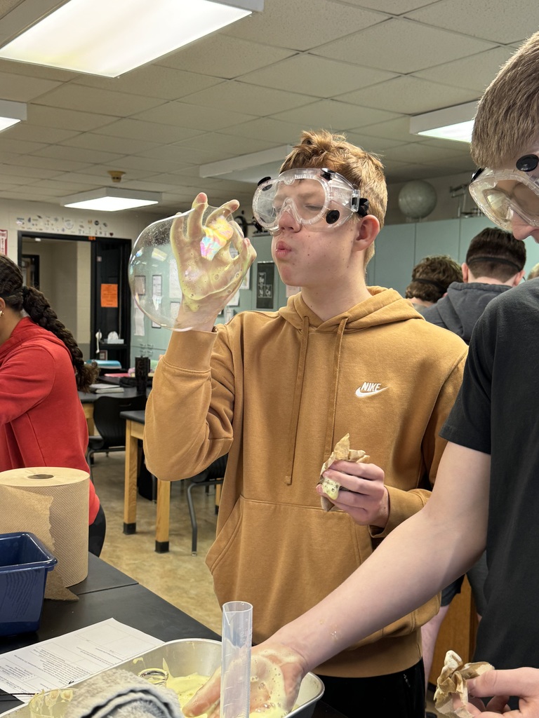 Student in safety goggles blows a large iridescent bubble formed from a foamy chemistry experiment.