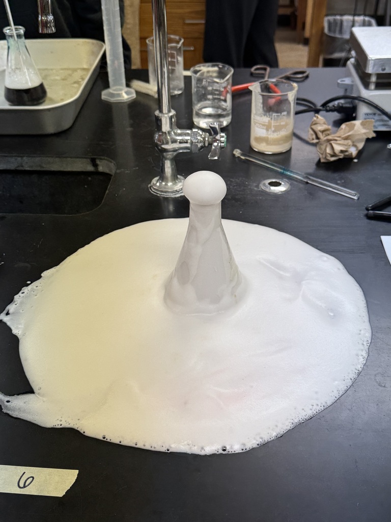 Close-up of a flask overflowing with white foam during a chemical reaction on a lab table.