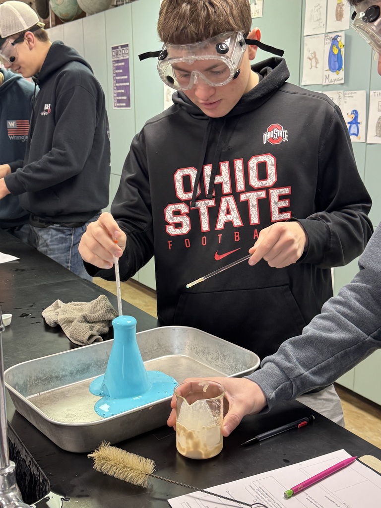 Students in safety goggles measure and pour liquids into flasks as foam rises during a lab experiment.