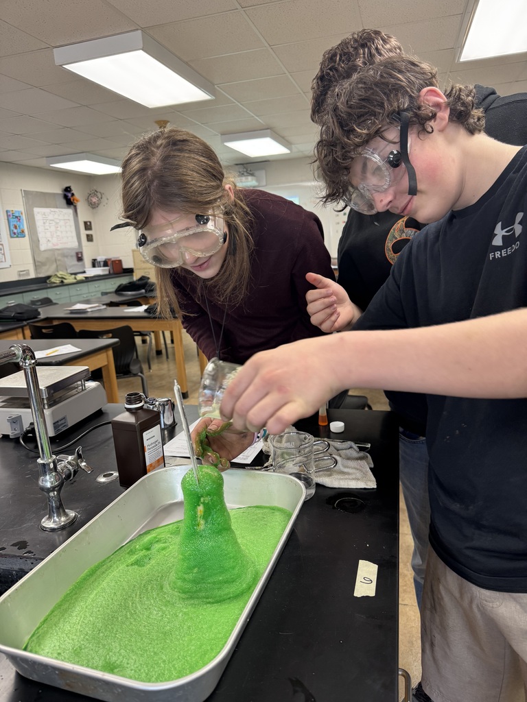 Two students wearing safety goggles conduct a chemistry experiment as green foam erupts from a flask in a lab tray.
