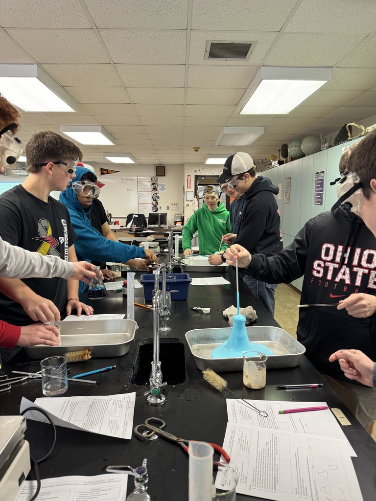 Group of students wearing safety goggles gathered around lab tables conducting foaming chemical reactions.