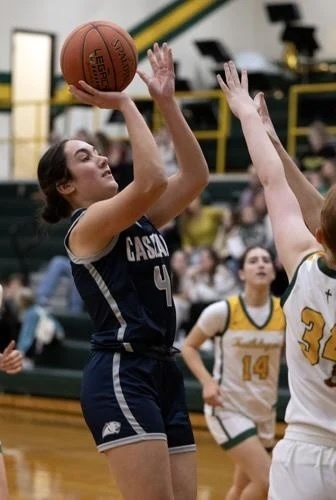 Girls basketball player in a dark Cascade uniform shoots a jump shot while a defender raises her arms to contest the play.