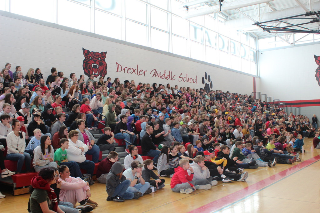 Large group of students and staff seated in bleachers inside a gymnasium with “Drexler Middle School” displayed on the wall.
