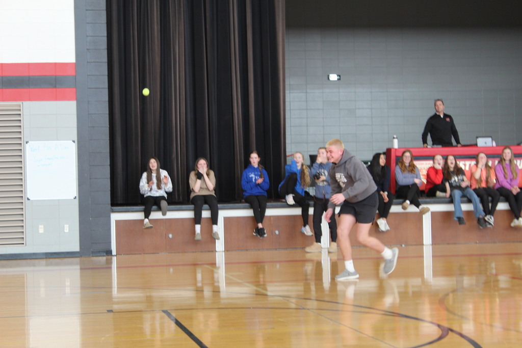 Student runs toward a tennis ball in a gym as other students sitting on the stage react and cheer.