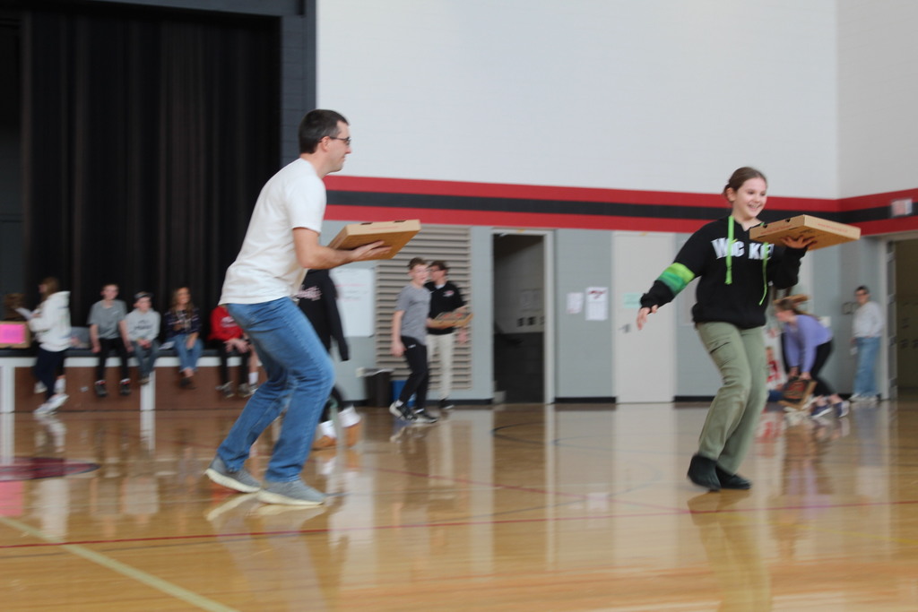 Students run across a gym floor holding pizza boxes during a school activity while classmates watch from the stage.