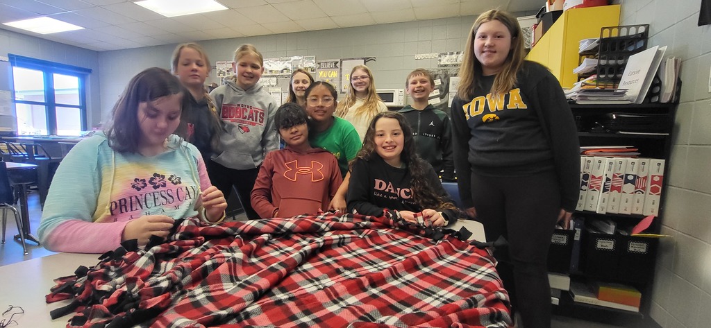 Group of students gathered around a table in a classroom working together to tie the edges of a red plaid fleece blanket.