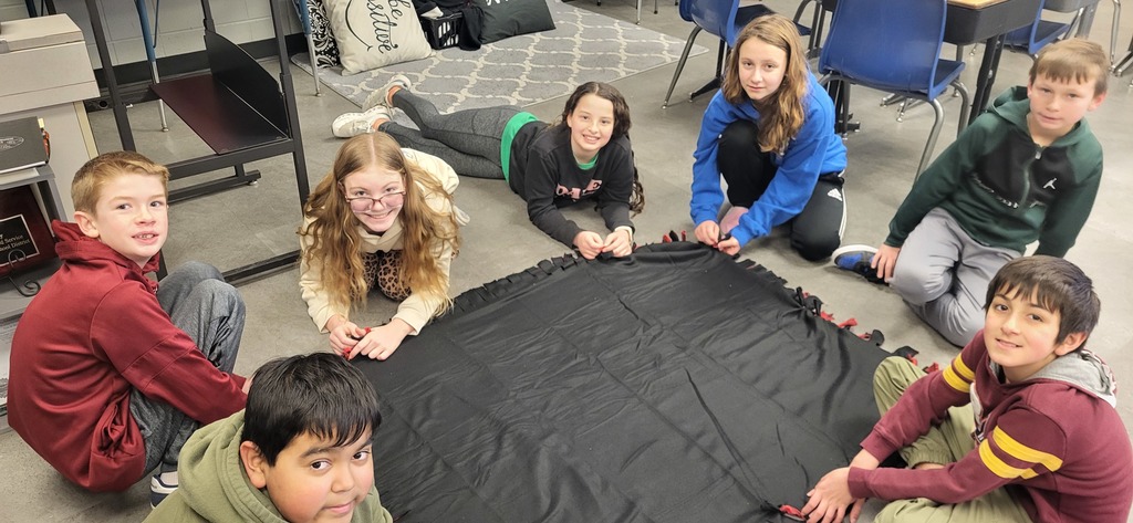 students sit on the classroom floor tying fabric strips around the edges of a black fleece blanket.