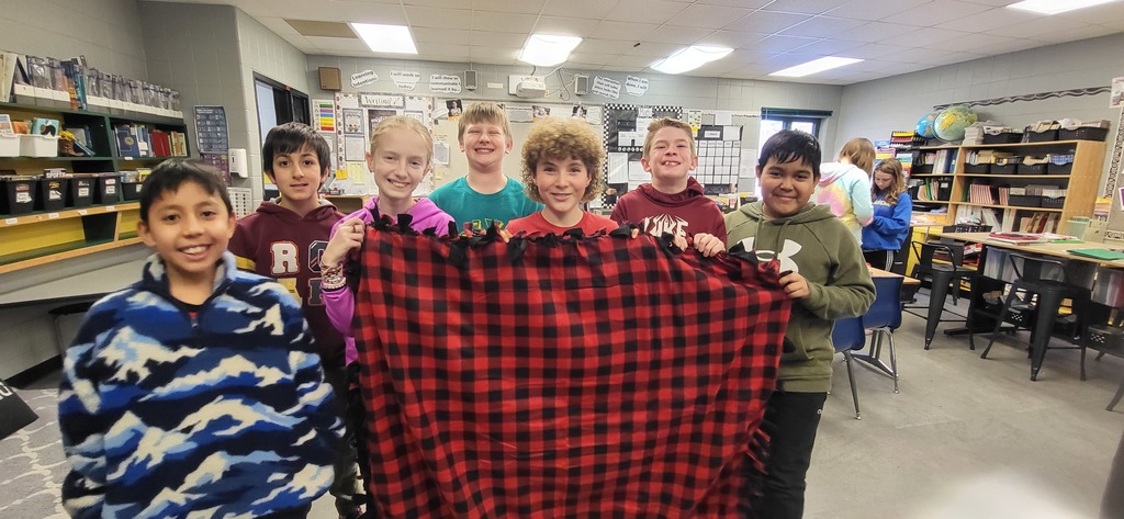Seven students stand in a classroom holding up a finished red and black plaid fleece blanket with tied edges.