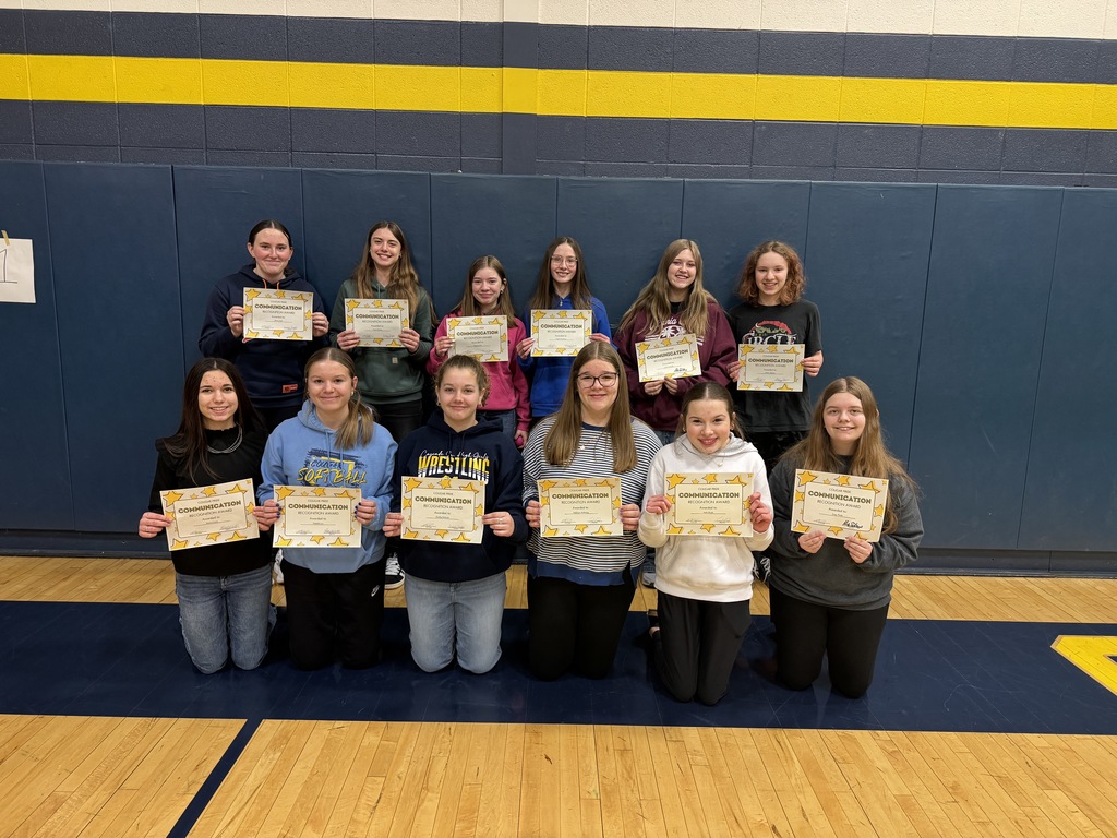 Group of students kneel and stand in a gym holding communication award certificates, smiling toward the camera.
