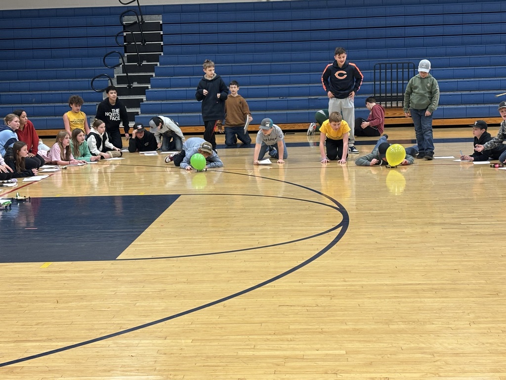 Students kneel on a gym floor blowing green and yellow balloons across the court while classmates watch from the sidelines.