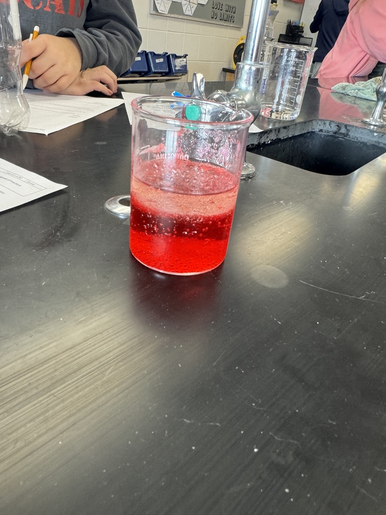 Beaker of bright red, bubbling liquid on a lab table, with students writing notes in the background.