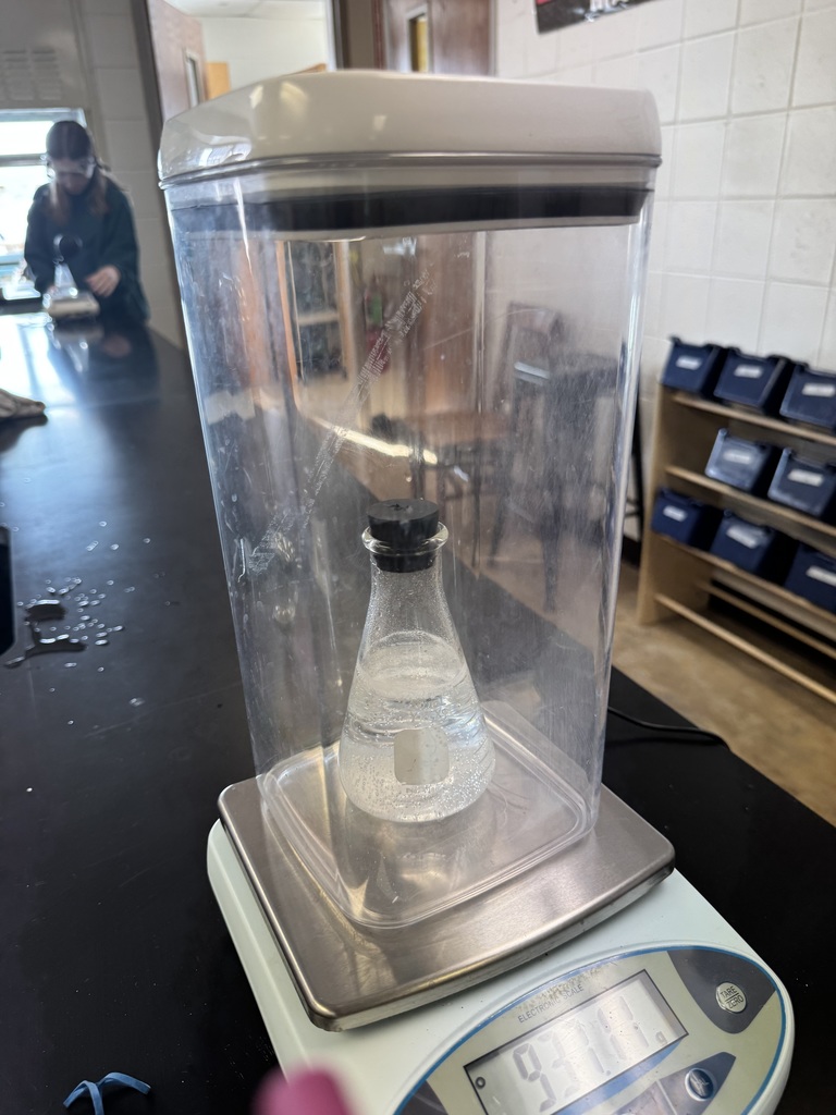 Stoppered flask with clear liquid sits on a digital scale inside a draft shield in a classroom lab.