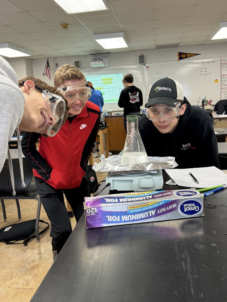 Three students wearing safety goggles lean toward a flask on a hot plate during a classroom experiment.