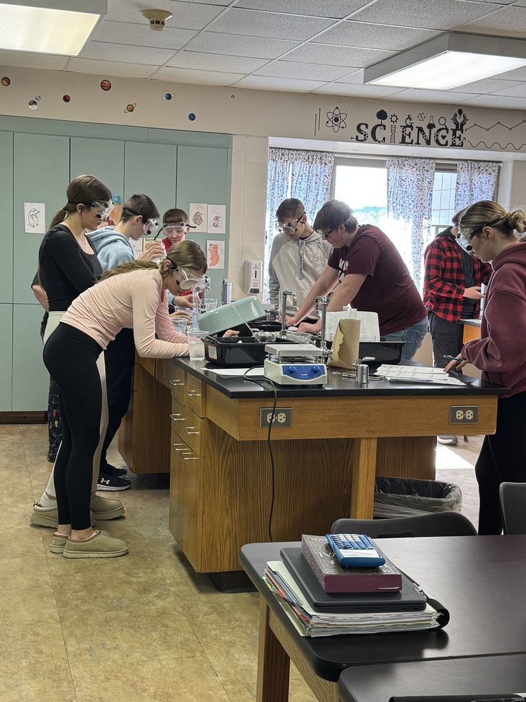 Students in safety goggles conduct a science lab experiment together at a classroom lab table.
