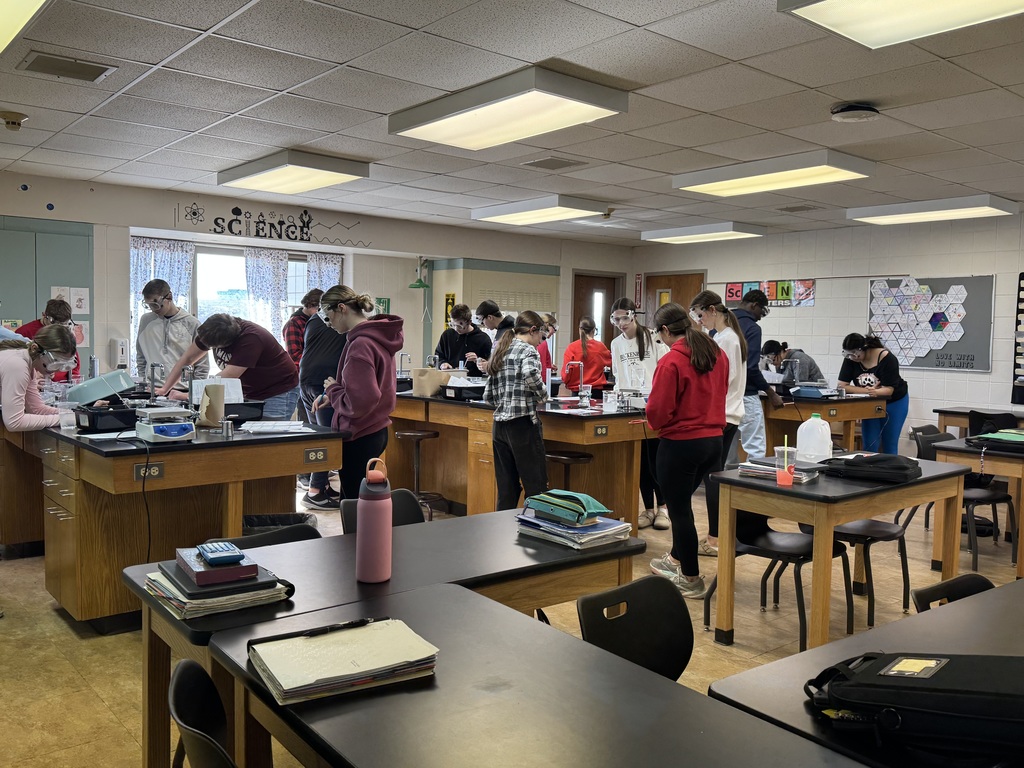 Students wearing safety goggles work in groups at lab stations during a science experiment in a classroom.