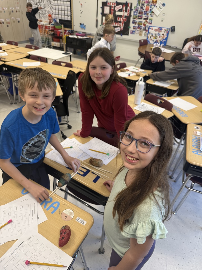 Three smiling students stand at desks with worksheets, potatoes, and sticks while working on a classroom experiment.