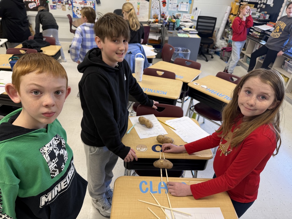 students stand at desks using potatoes and wooden sticks to test balance while working on a classroom activity sheet.