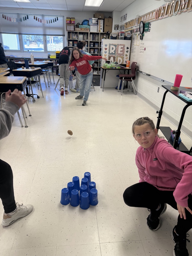 Student rolls a potato toward a pyramid of blue cups while classmates watch and another student crouches near the target.