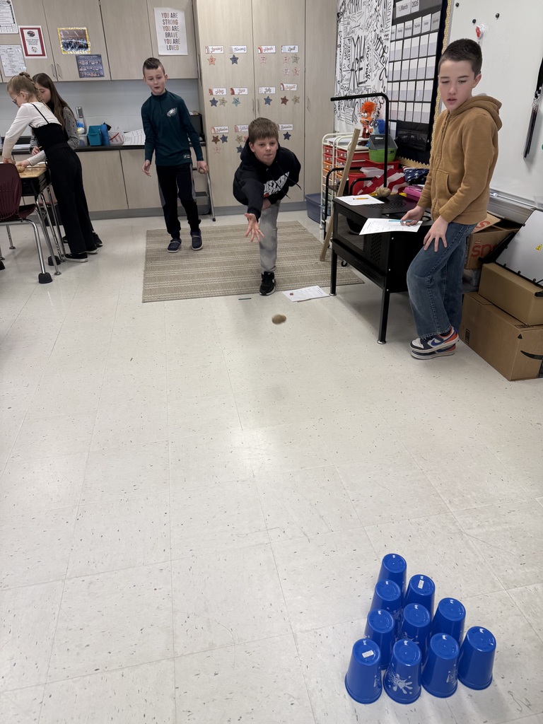 Student tosses a potato toward stacked blue cups on the floor as classmates observe during a hands-on classroom activity.
