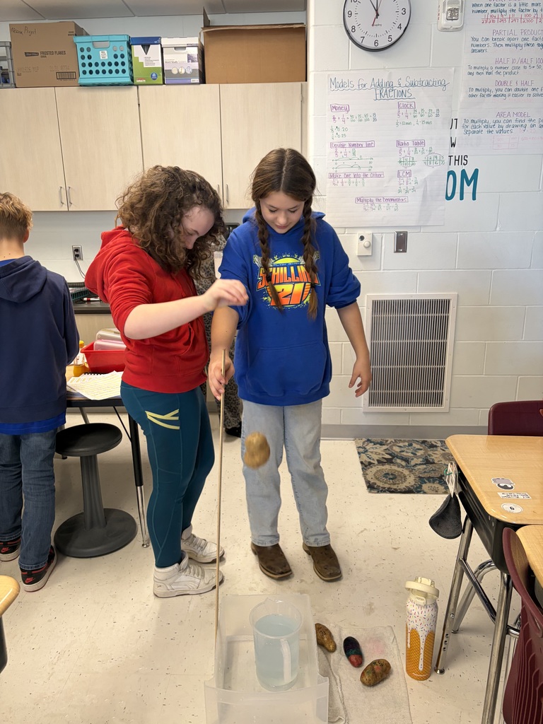 Two students use a stick to lower a potato toward a container of water during a hands-on classroom science activity.