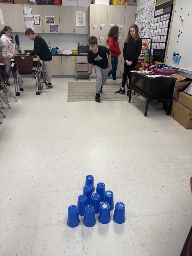 Student rolls a potato across the classroom floor toward a triangle of blue plastic cups while classmates watch nearby.