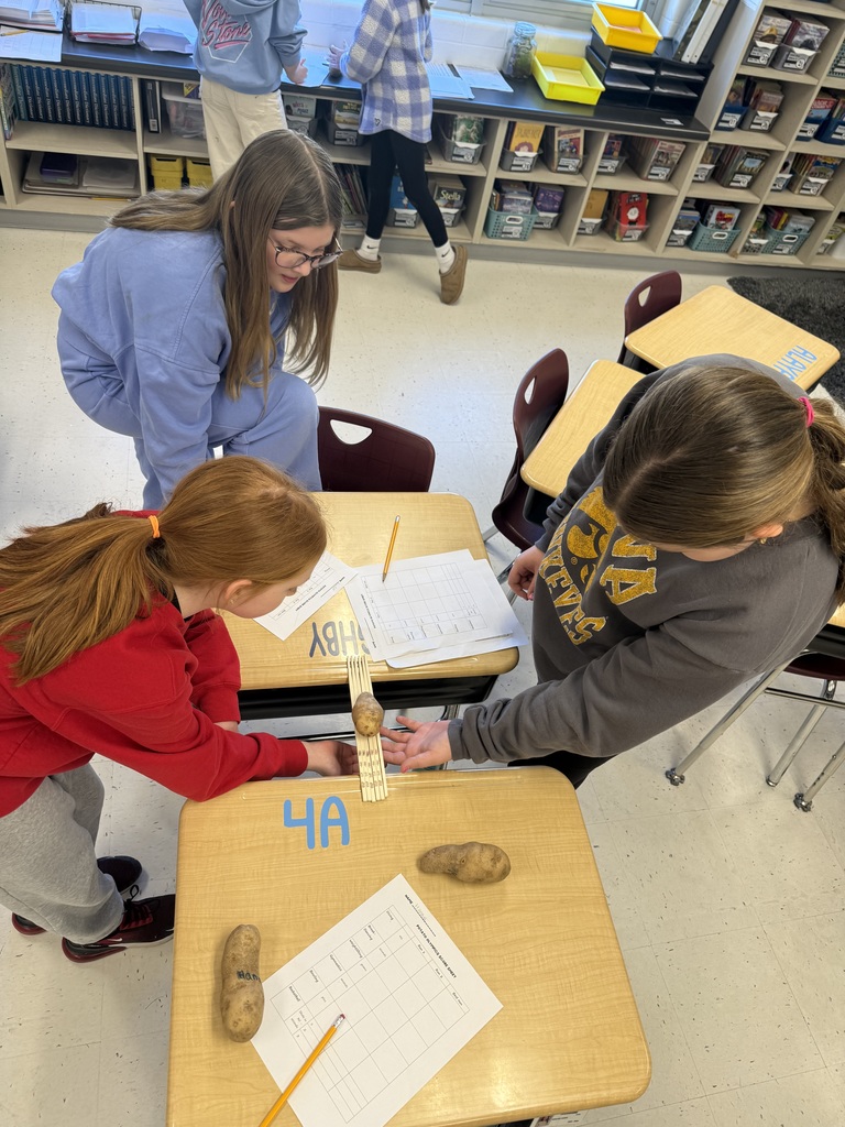 Three students collaborate at desks, balancing a potato on wooden sticks while recording results on a worksheet.