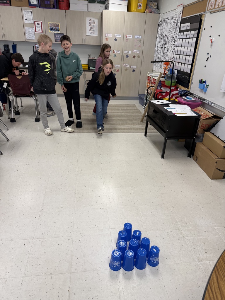 Student rolls a potato toward stacked blue cups on the floor while classmates stand nearby waiting their turn.