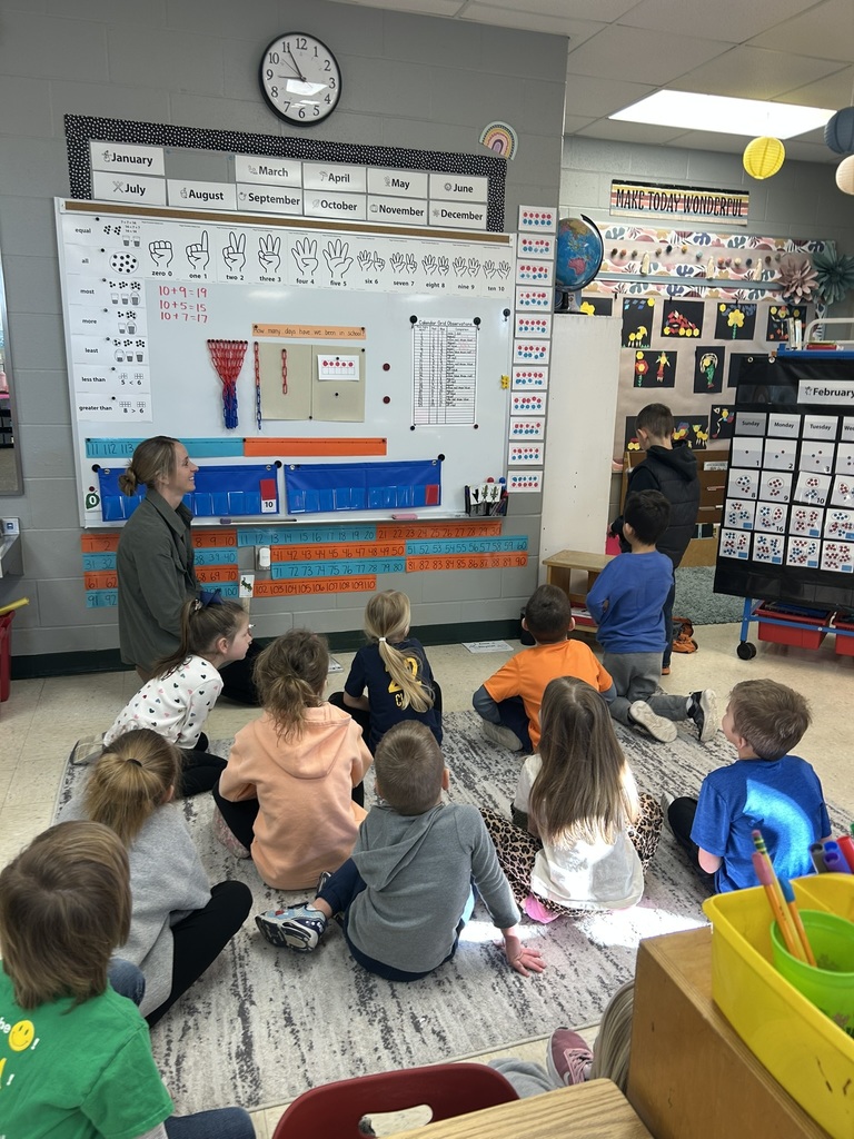 Students sit on a rug facing a teacher who leads a calendar and math routine using a classroom wall display.