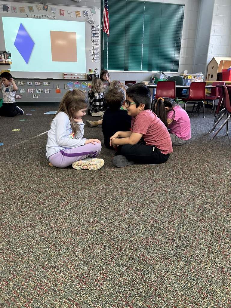 Small groups of students sit on the carpet talking and working together while shapes are projected on the classroom board.