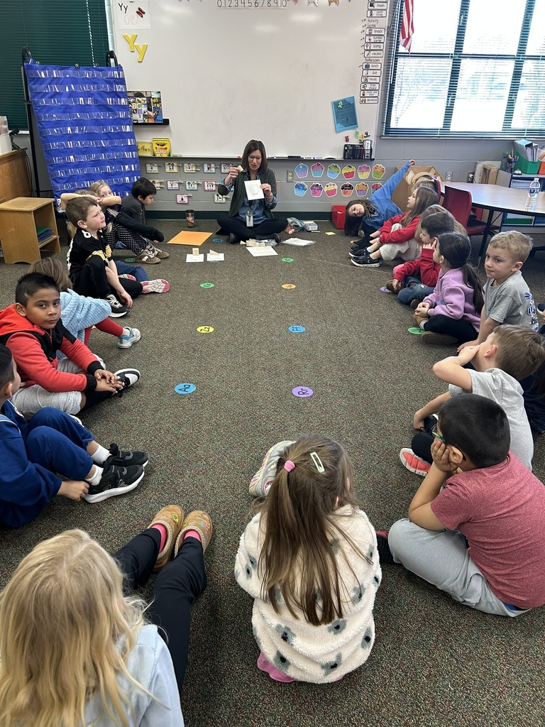 Teacher sits with students in a circle on the carpet while holding a card and leading a group learning activity.