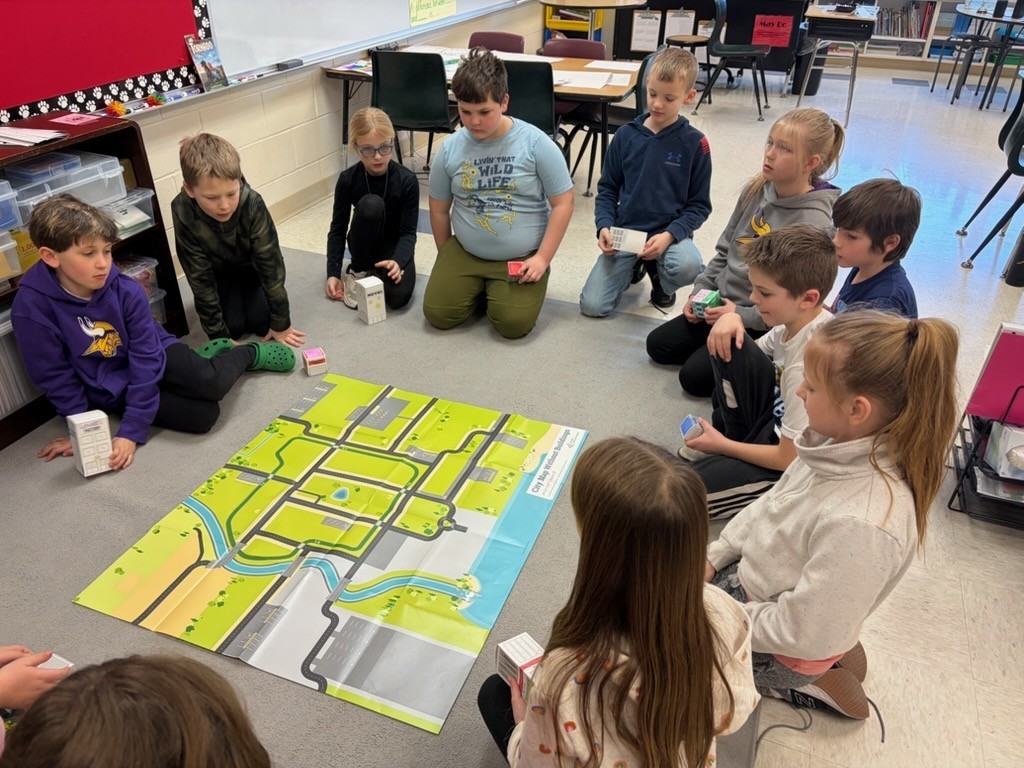 Students sit in a circle around a large map mat while holding small building blocks during a classroom planning activity.