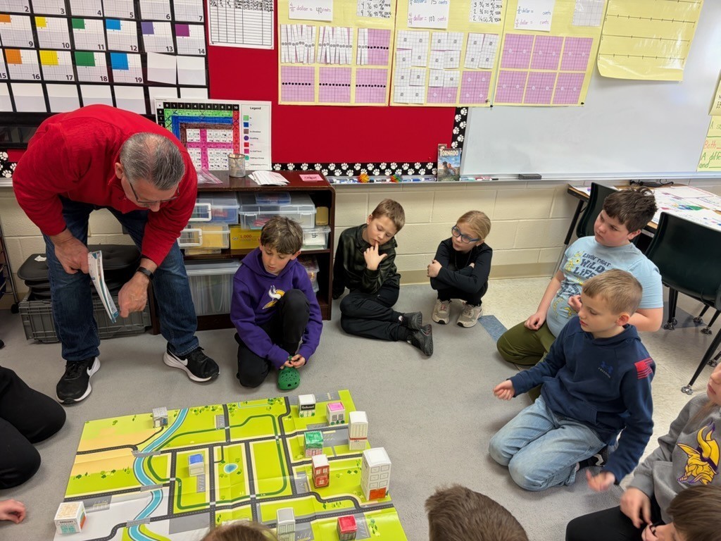 Teacher kneels beside students gathered around a city map mat while discussing where to place buildings in a classroom activity.