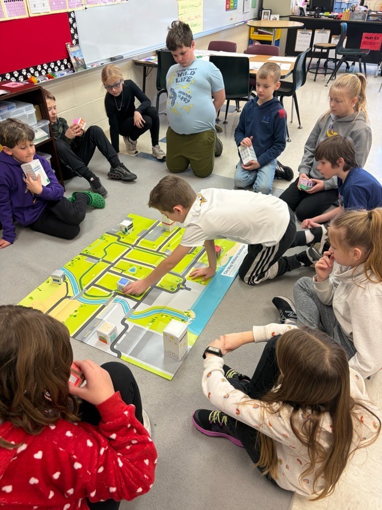 Student places a small building block on a city map mat while classmates watch and plan together.