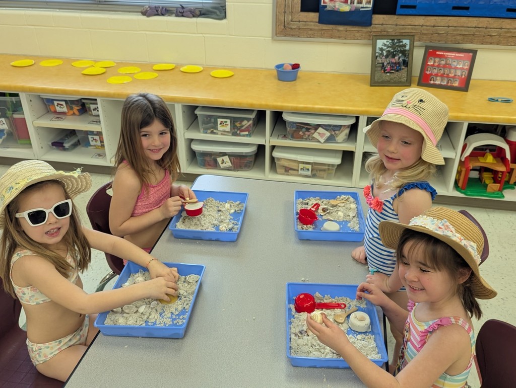 Four preschool students sit at a table exploring sand trays with shells and scoops while wearing beach hats and summer clothes.