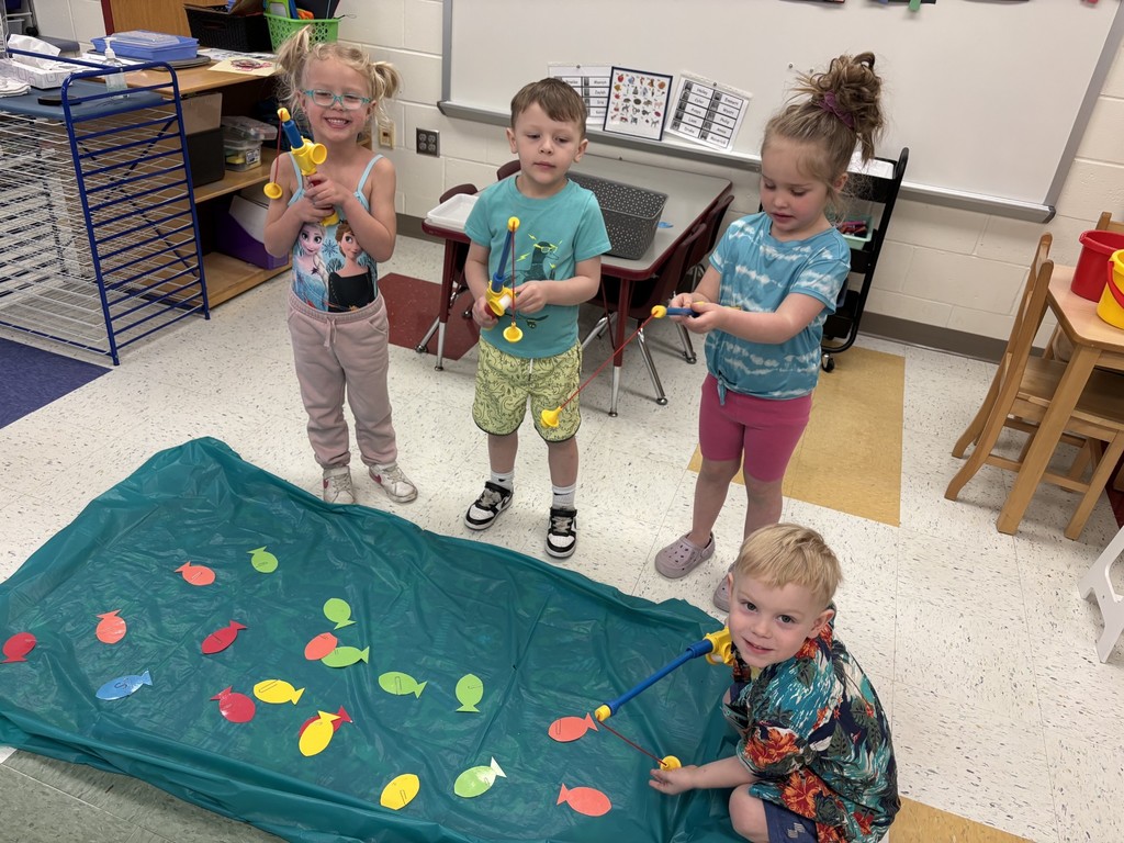 Preschool students use toy fishing rods to catch colorful paper fish from a small play pond during a classroom game.