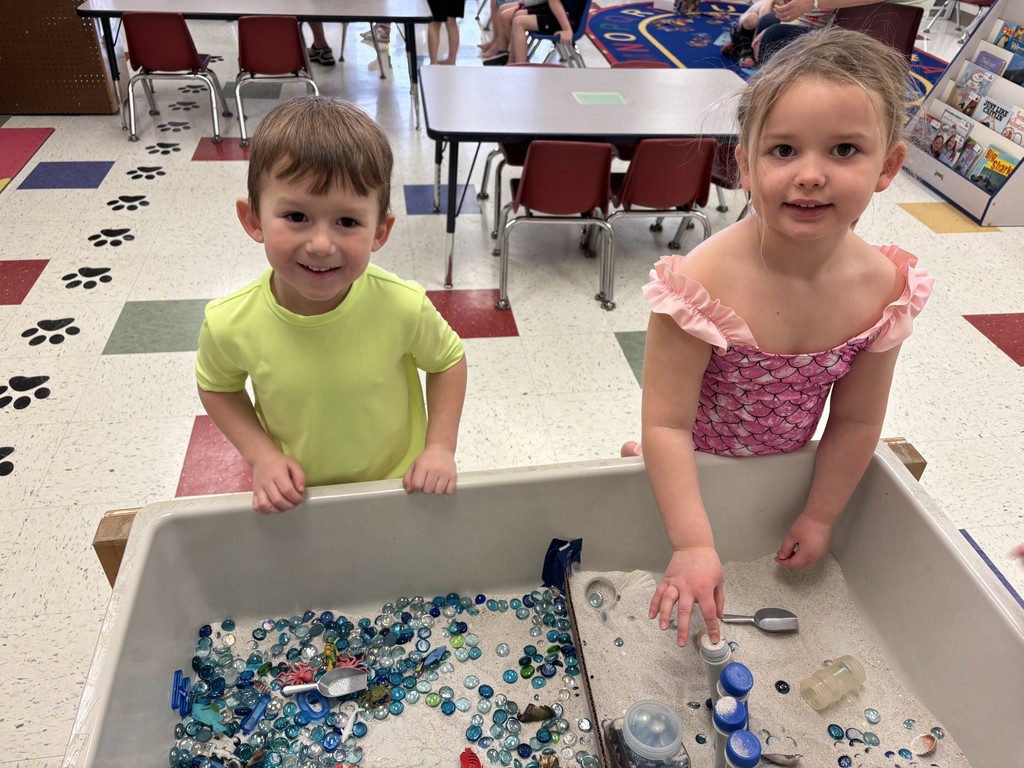 students wearing summer hats and swimwear play with water and toy sea animals at a sensory table.