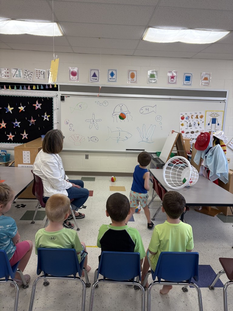 Students sit facing a whiteboard with ocean drawings while one student tosses a small ball during a classroom counting activity.