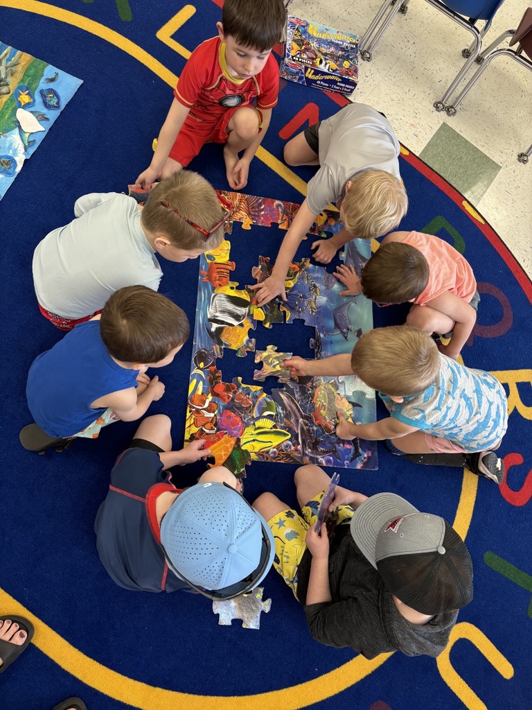 A group of young students sit together on a carpet working cooperatively to assemble an ocean-themed puzzle.