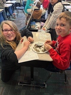 Two students smile at the camera while sorting through soil on a plate during a classroom science activity.