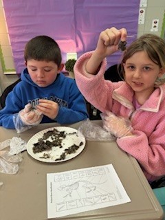 Two students wearing gloves examine soil samples on a plate while completing a science worksheet at their desk.