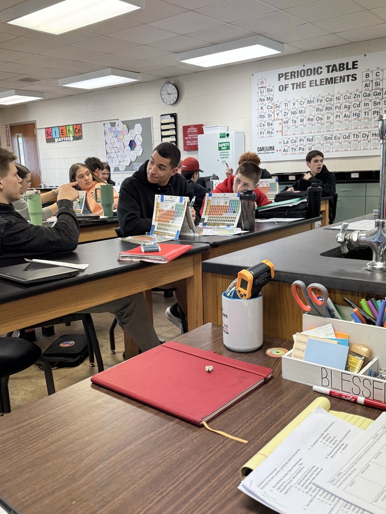 Students sit at lab tables in a science classroom while discussing and referencing periodic table charts during a lesson.