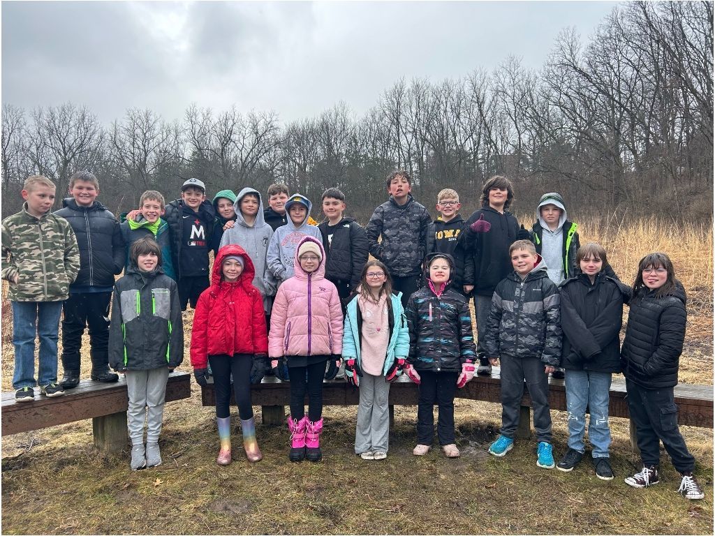 Group of students stand outdoors on a wooden platform, bundled in jackets, posing together in a grassy area with leafless trees.